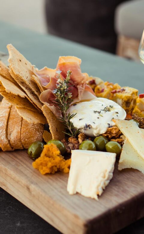 charcuterie board with breads, crackers, meats and cheeses next to two glasses of white wine